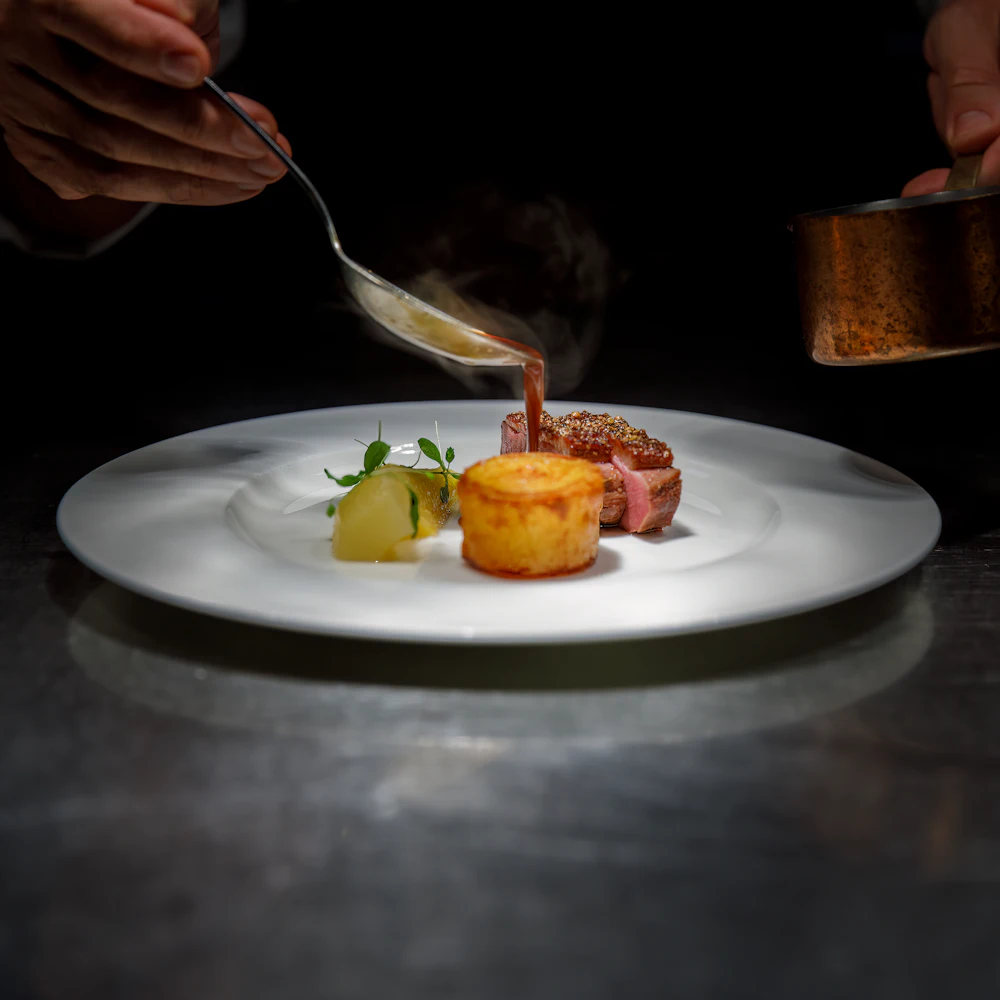 Sauce wird mit einem Löffel über ein angerichtetes Fleischgericht mit Kartoffeltörtchen gegossen